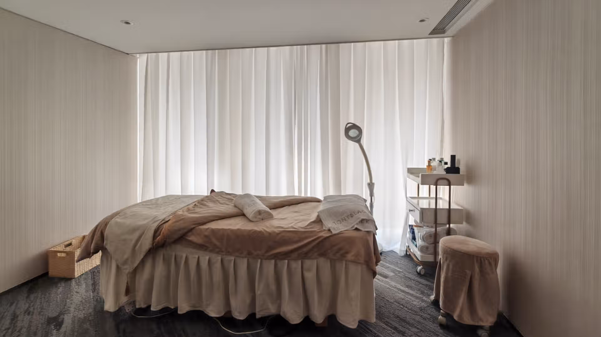 香港美容院裝修工程：高檔美容診療室（Treatment Room）空間規劃，展示配有粉色美容床與白色落地紗窗布藝軟裝的極簡奶油風房間，兩側牆面採用淺米色垂直紋理壁紙飾面
