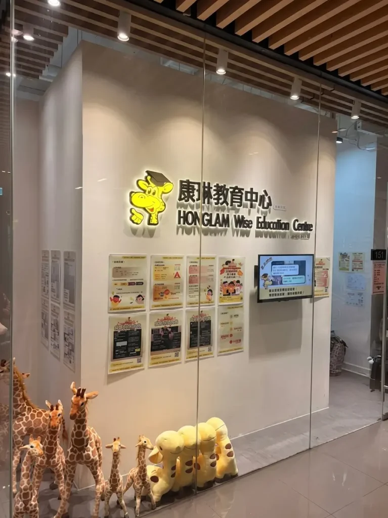 商業空間門面裝修公司：兒童教育中心前台與宣傳牆規劃，暖白色飾面背牆安裝LED背發光金屬字招牌與液晶顯示屏，玻璃幕牆前設有玩偶展示區，上方配備木質條狀格柵天花與軌道射燈